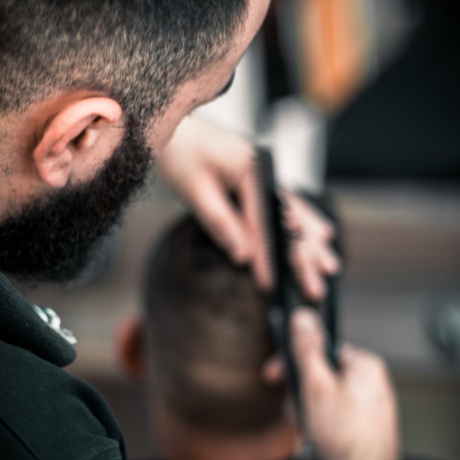 Barbeiro de barba escura apara o cabelo de um cliente sentado na cadeira da barbearia, segurando pente e máquina de corte
