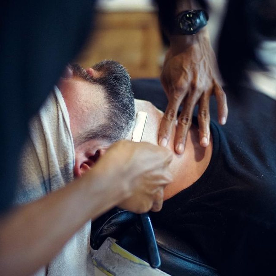 Cliente reclinado na cadeira da barbearia, com toalha no pescoço, enquanto o barbeiro acerta a barba com navalha