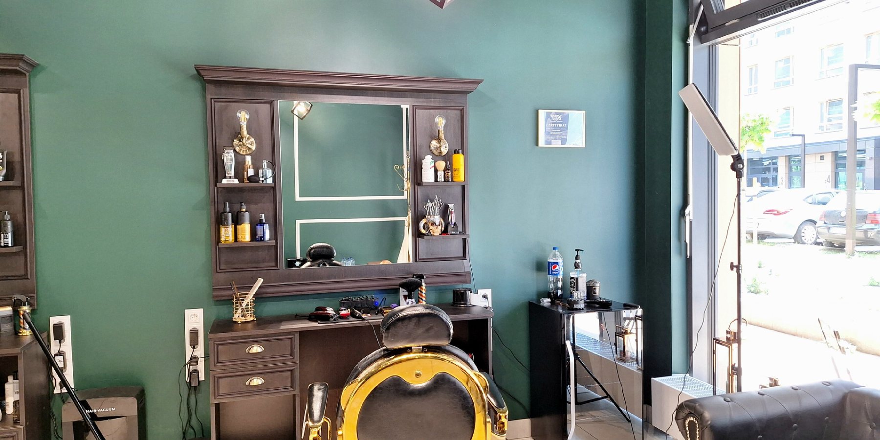 Interior de barbearia com parede verde-escura, bancada de madeira com espelho emoldurado e luminárias, produtos e máquinas nas prateleiras, cadeira de barbeiro dourada em frente ao espelho e luz natural entrando pela vitrine aberta para a rua