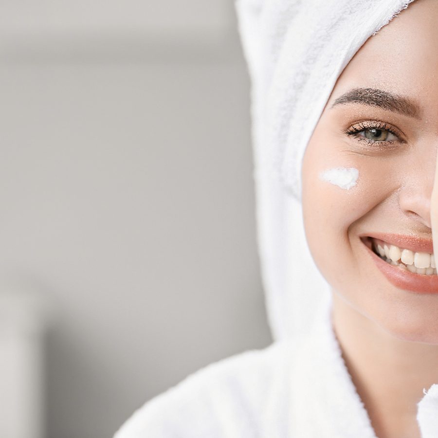 Mulher sorridente de roupão branco e toalha na cabeça com creme facial aplicado na bochecha, em ambiente claro de clínica de estética