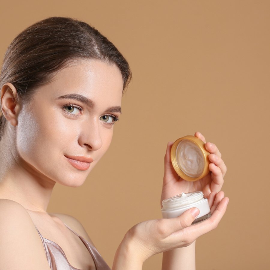 Mulher de pele luminosa segurando dois potes abertos de creme facial, um com tampa dourada e outro branco, sobre fundo bege claro de estúdio