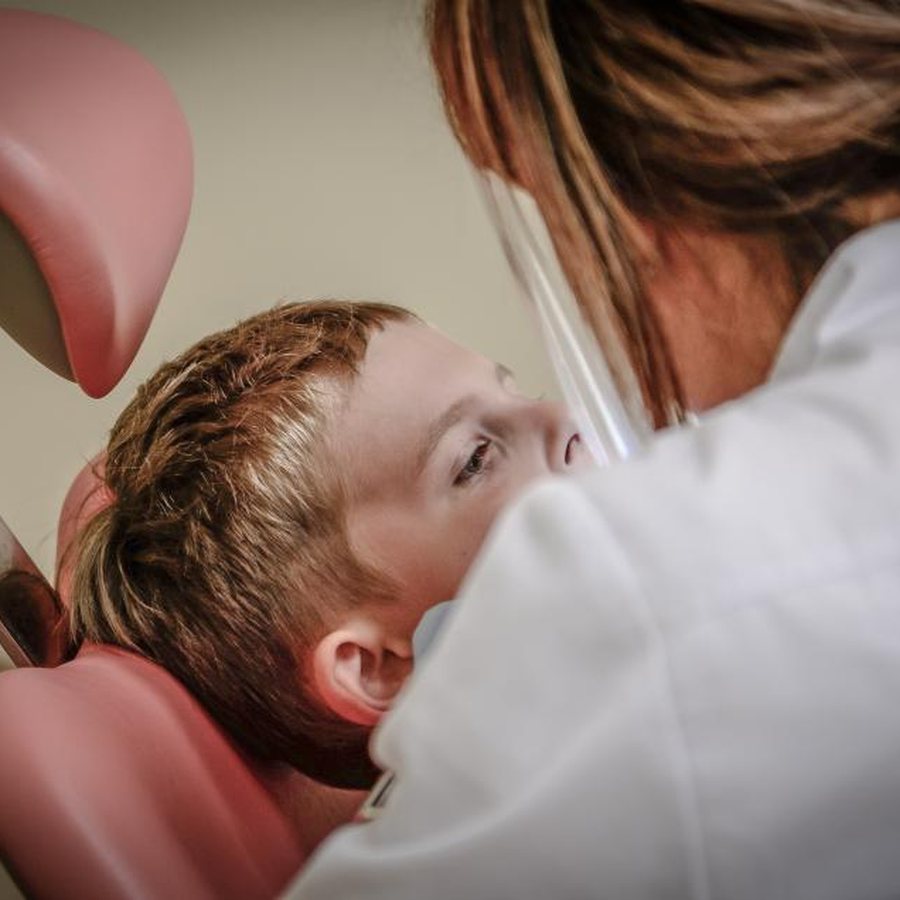 Dentista de jaleco branco e protetor facial inclinada sobre um menino deitado na cadeira odontológica rosa, examinando os dentes dele de perto