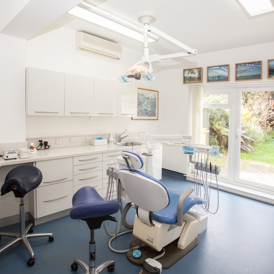 Sala de atendimento odontológico ampla e clara, com cadeira azul ao centro, dois mochos, refletor de teto, bancada e armários brancos e uma porta de vidro que dá para um jardim iluminado pelo sol