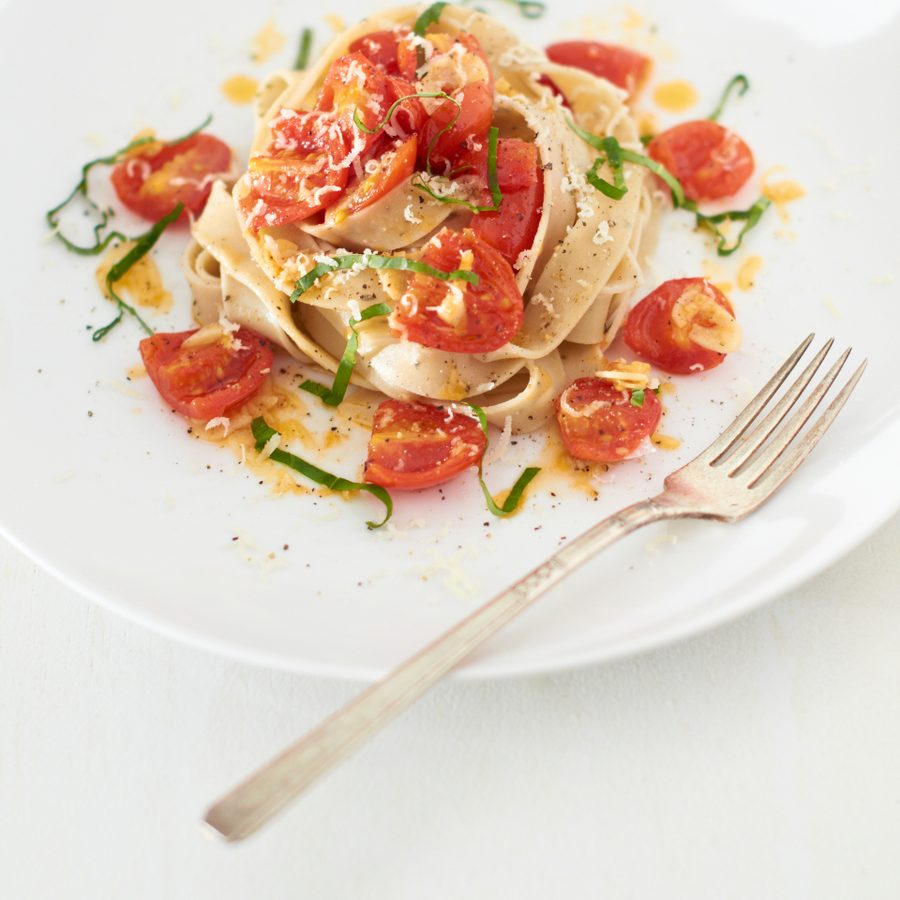 Prato de fettuccine com tomates-cereja, manjericão e queijo ralado servido em prato branco com garfo ao lado