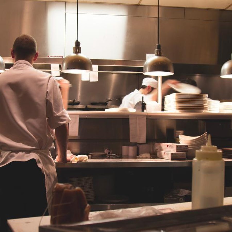 Cozinheiros preparando pratos na cozinha profissional do restaurante, com bancada de inox e lâmpadas de aquecimento sobre o passe