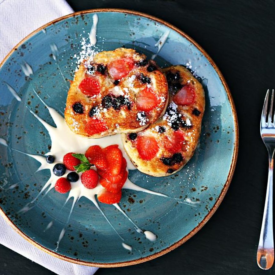 Sobremesa de panquecas com frutas vermelhas, creme e açúcar de confeiteiro servida em prato azul sobre mesa escura