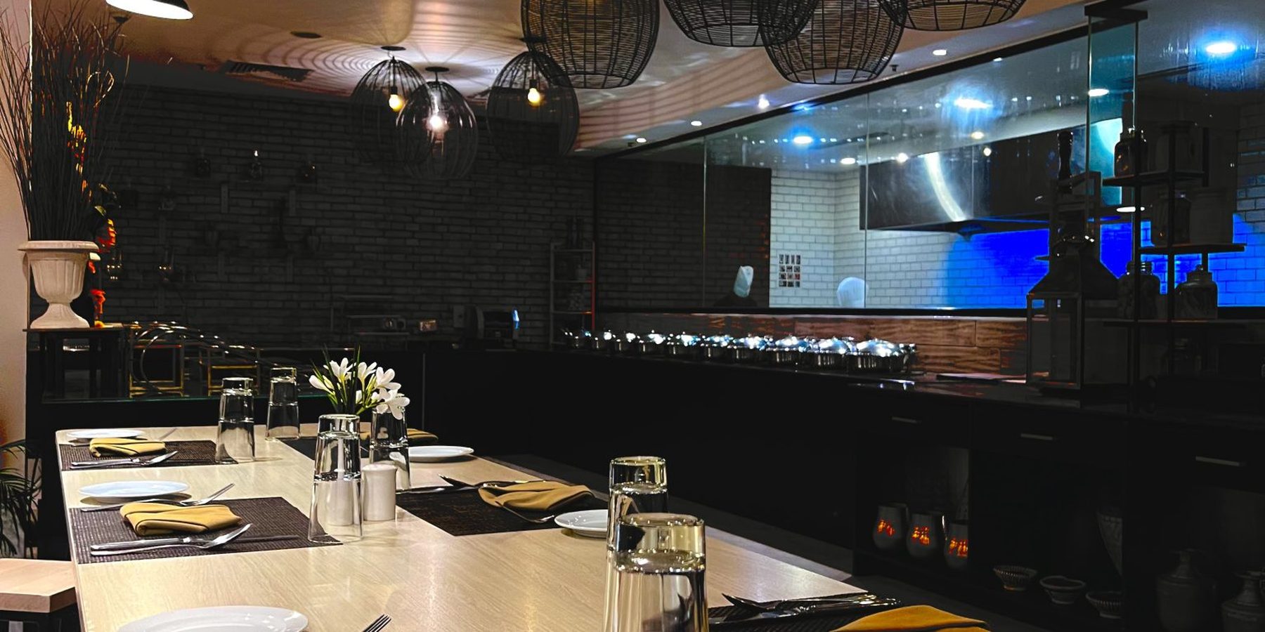 Mesa comprida posta com pratos, taças e guardanapos no salão do restaurante, com luminárias pendentes e cozinha aberta ao fundo